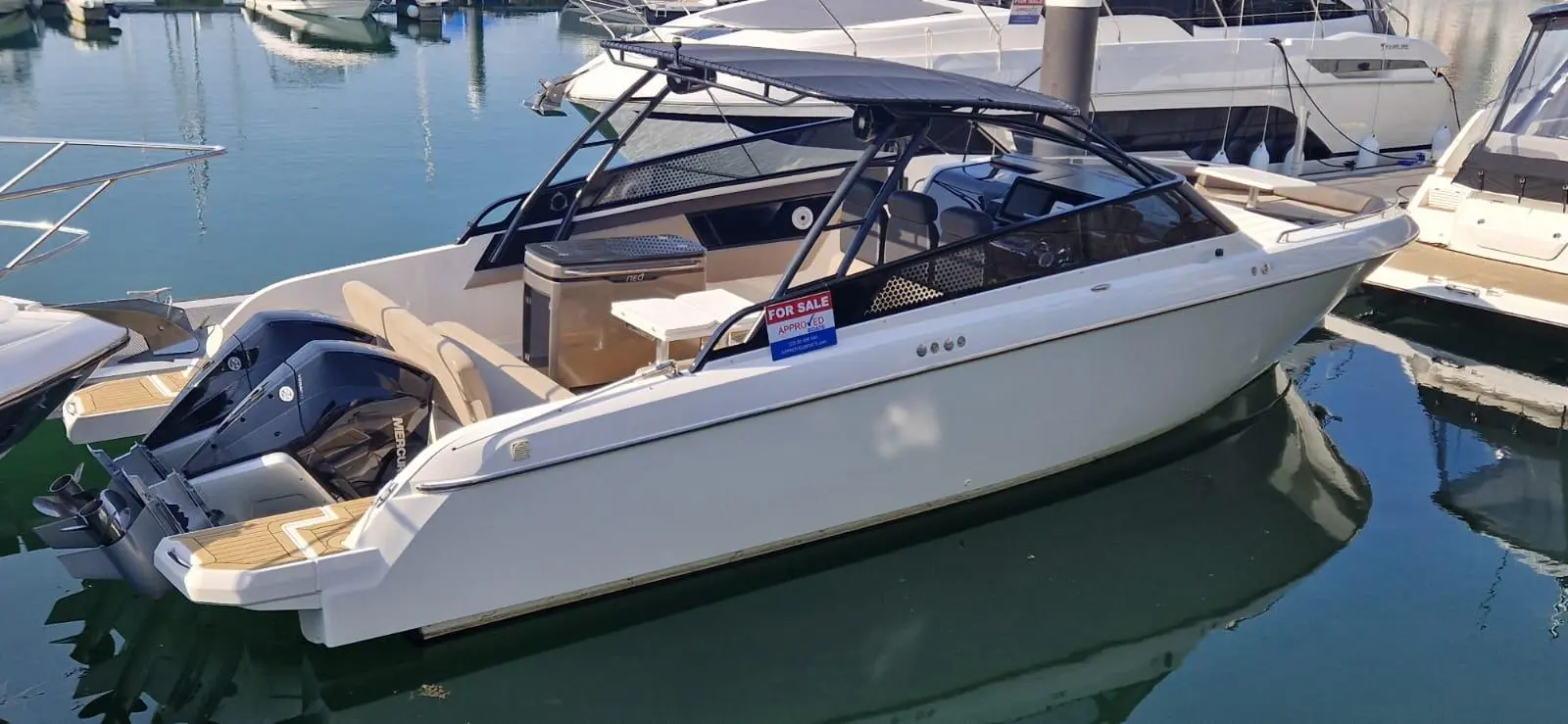White motorboat docked in a marina with a "FOR SALE" sign visible on the side and luxury boats in the background.